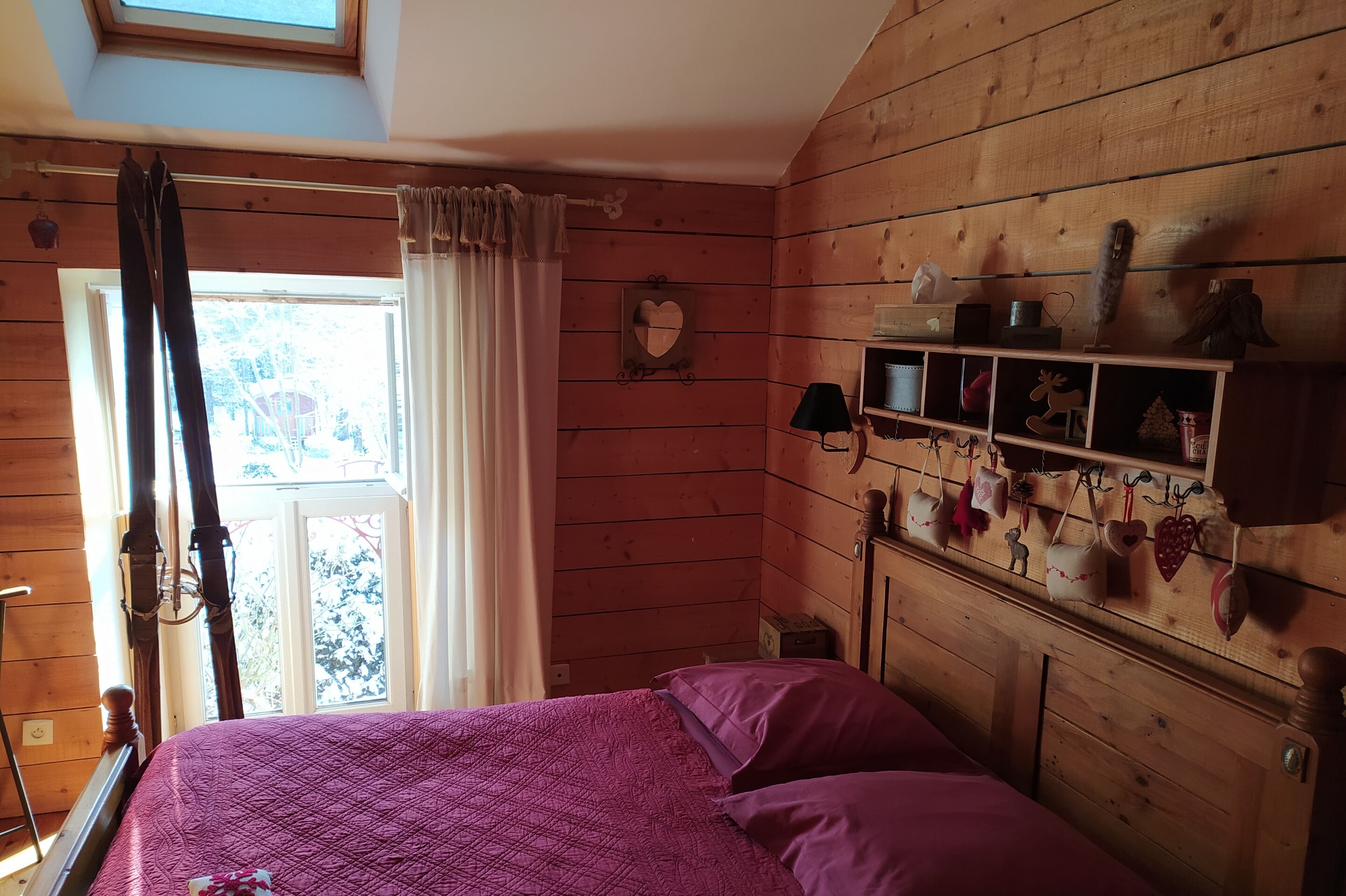 Ein Zimmer aus Holz mit einem Bett, einem Fenster mit Vorhängen und einem Regal mit herzförmigen Dekorationen.