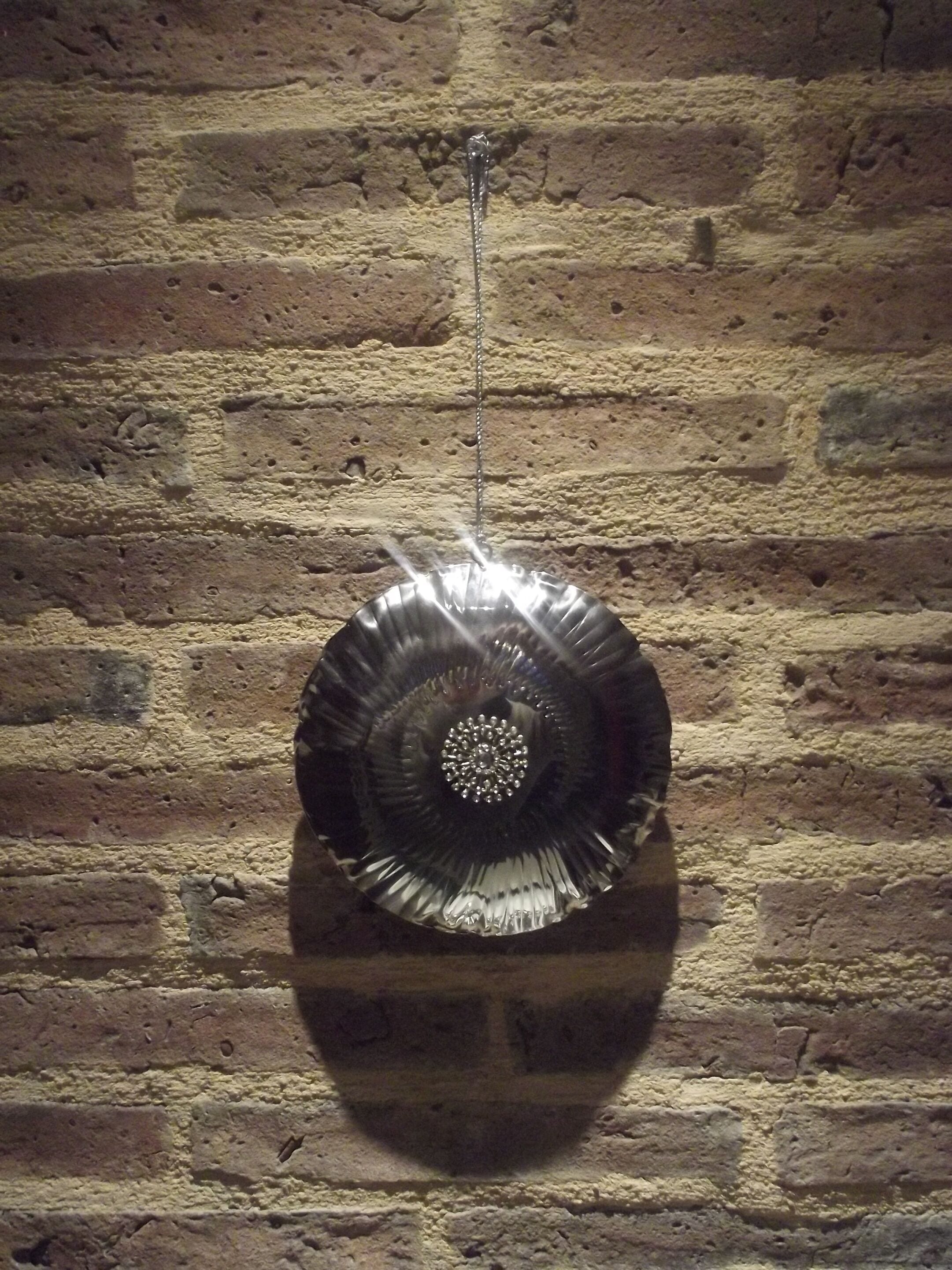 Un objet métallique, de forme circulaire, orné d’un motif central, suspendu contre un mur de briques.