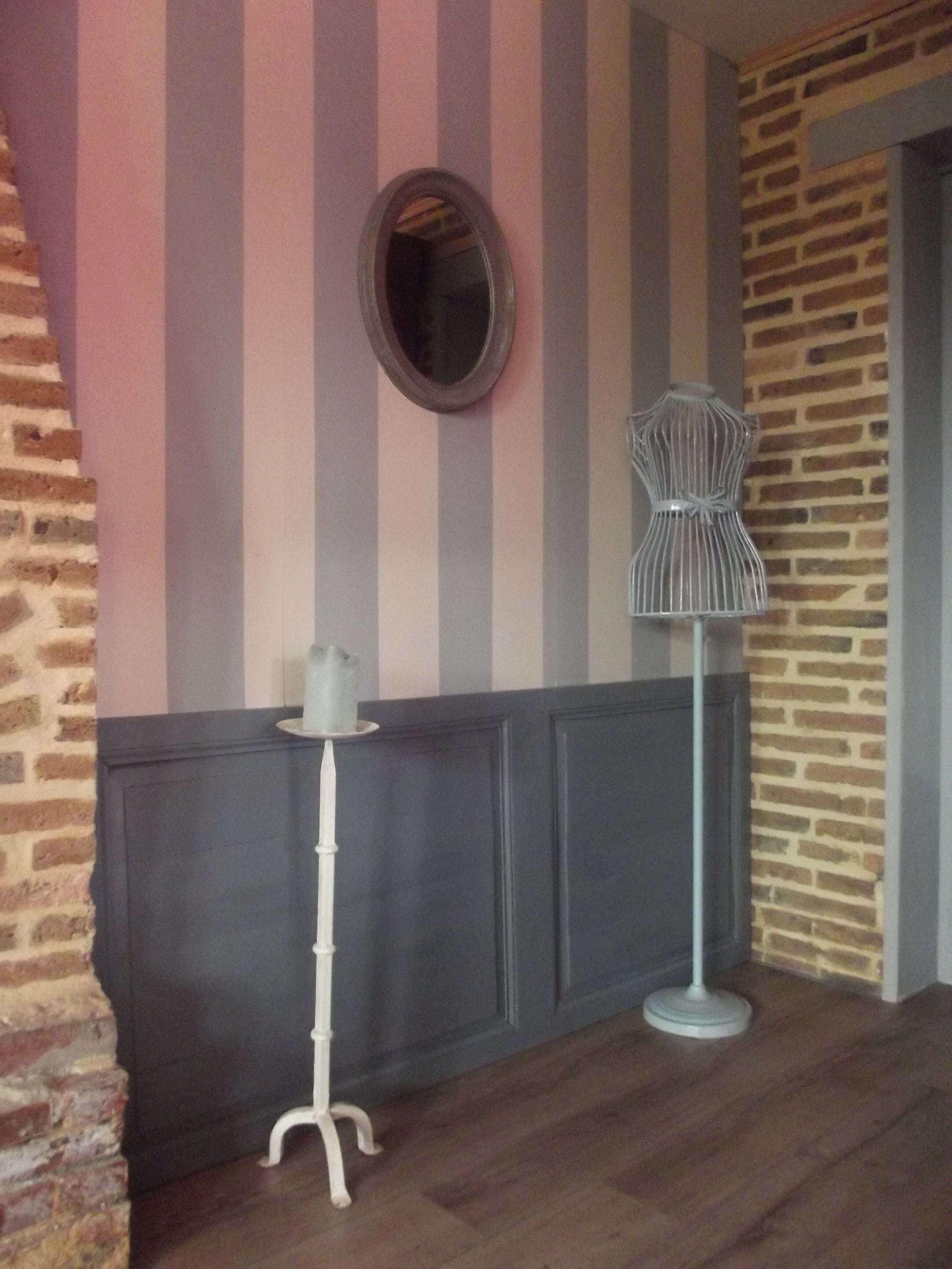 A corner with a striped wall, a round mirror, a white pedestal table, and a wireframe chair.
