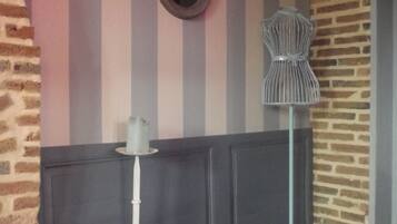 A corner with a striped wall, a round mirror, a white pedestal table, and a wireframe chair.