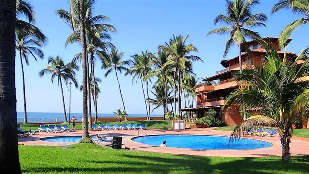 A resort pool area with multiple lounge chairs, palm trees, and a building with a red roof. - Unlimited Luxury Villa  LOS TULES (Puerto Vallarta)