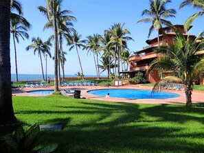A resort pool area with multiple lounge chairs, palm trees, and a building with a red roof. - Unlimited Luxury Villa  LOS TULES (Puerto Vallarta)