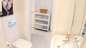 Une petite salle de bain comprenant des toilettes, un lavabo et un miroir. On y trouve un panier sur une étagère et une affiche accrochée au mur.