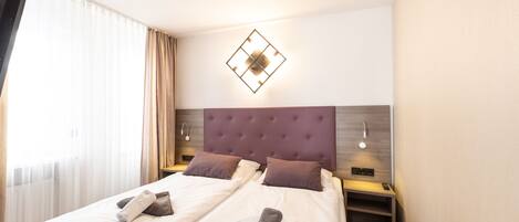 Une chambre d’hôtel avec deux lits, une tête de lit agrémentée d’un luminaire aux motifs géométriques, des tables de chevet avec des lampes et une fenêtre avec des rideaux.