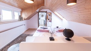 Un dormitorio con techo inclinado, vigas de madera, una cama con almohadillas, una mesita de noche con lámpara y una ventana con vistas.