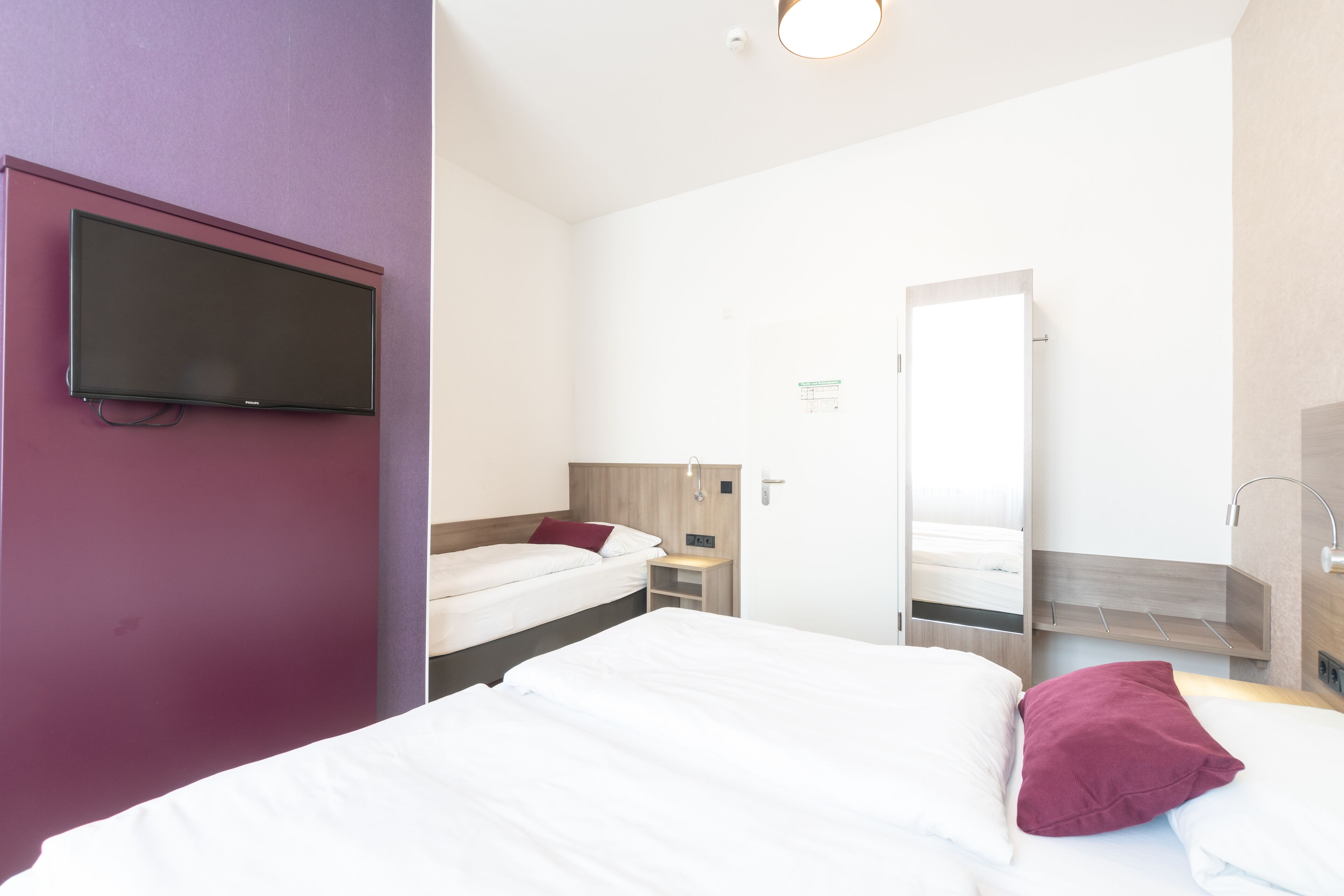 Ein Hotelzimmer mit einem großen Bett, einem kleineren Bett, einem an der Wand befestigten Fernseher, einem Nachttisch und einem Spiegel.