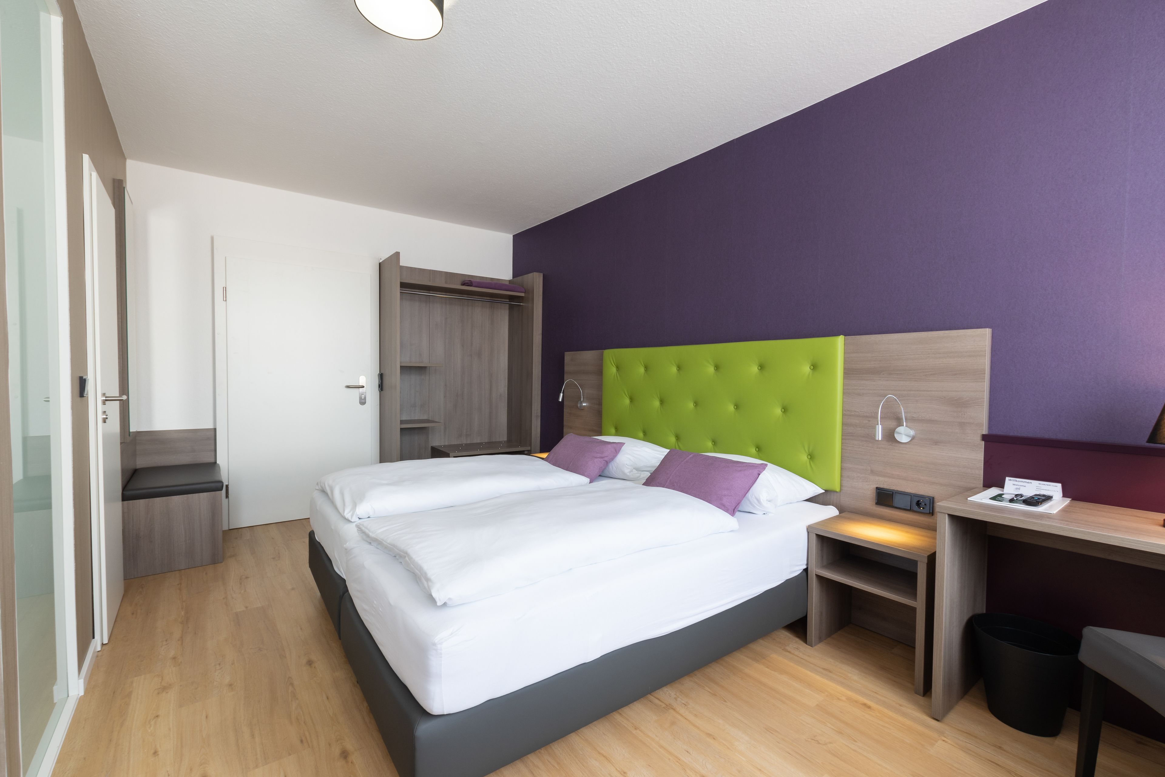 A modern hotel room with a large bed, a desk, and a wardrobe.