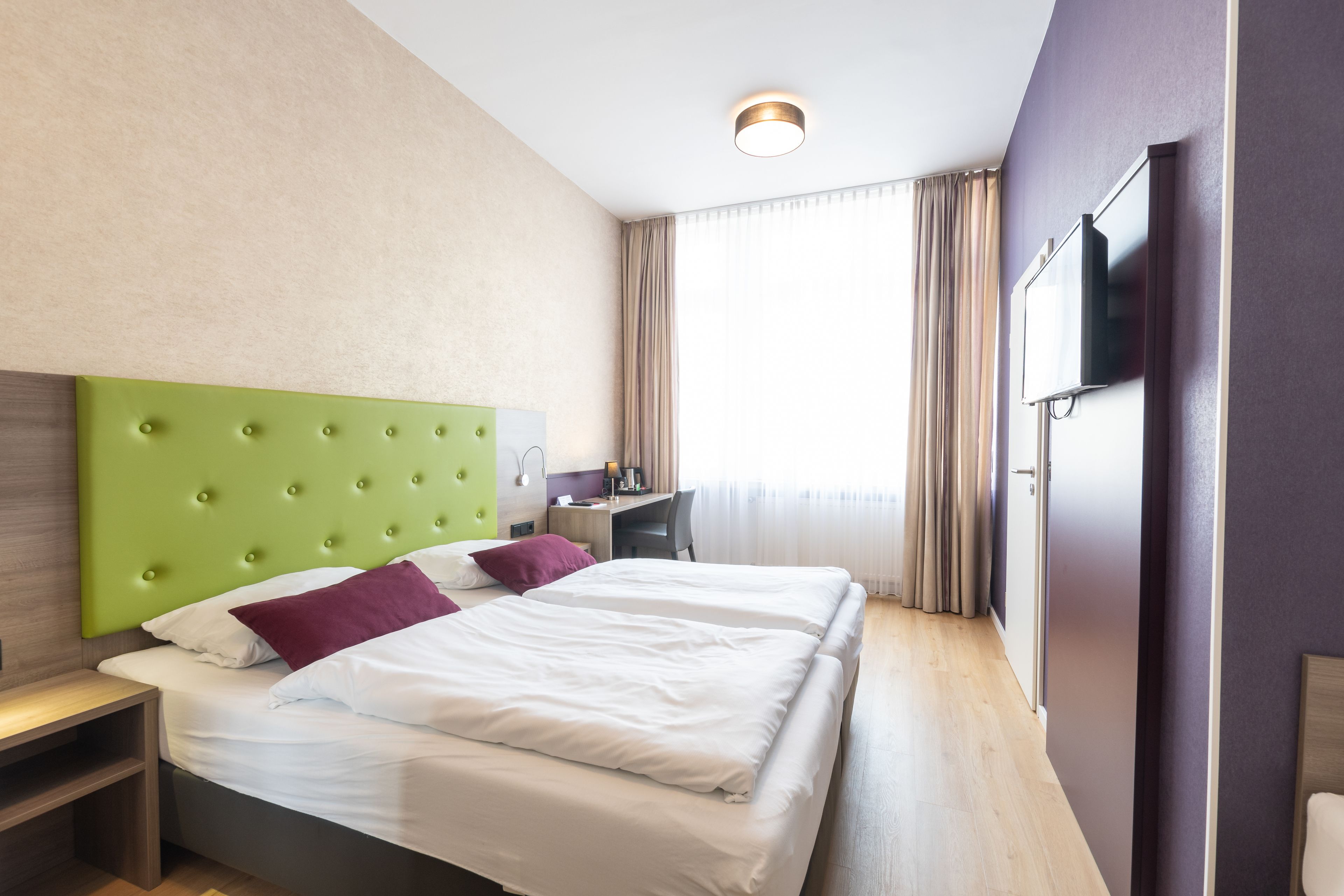 Habitación de hotel con cabecera verde, dos camas con sábanas blancas y almohadas moradas, un escritorio con silla, un televisor de pantalla plana en la pared y un ventanal con cortinas.