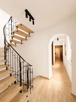 Una escalera de caracol con peldaños de madera y barandillas de metal negro, que conduce a un pasillo con suelo de madera y paredes blancas.