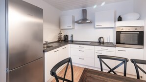 Una cocina moderna con armarios blancos, un refrigerador de acero inoxidable, una estufa a gas y un microondas. Hay dos sillas negras alrededor de una mesa de madera.
