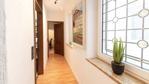 Un pasillo con piso de madera, una maceta con planta de aloe vera, un cuadro enmarcado y una ventana con diseño de cuadrícula.