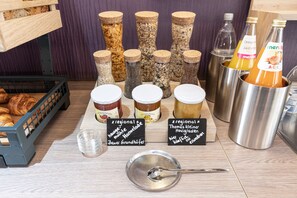 A variety of food items and condiments on a wooden table, including jars of preserves, bottles of orange juice, and a basket of croissants. - Hotel Porta Nigra (Trier)
