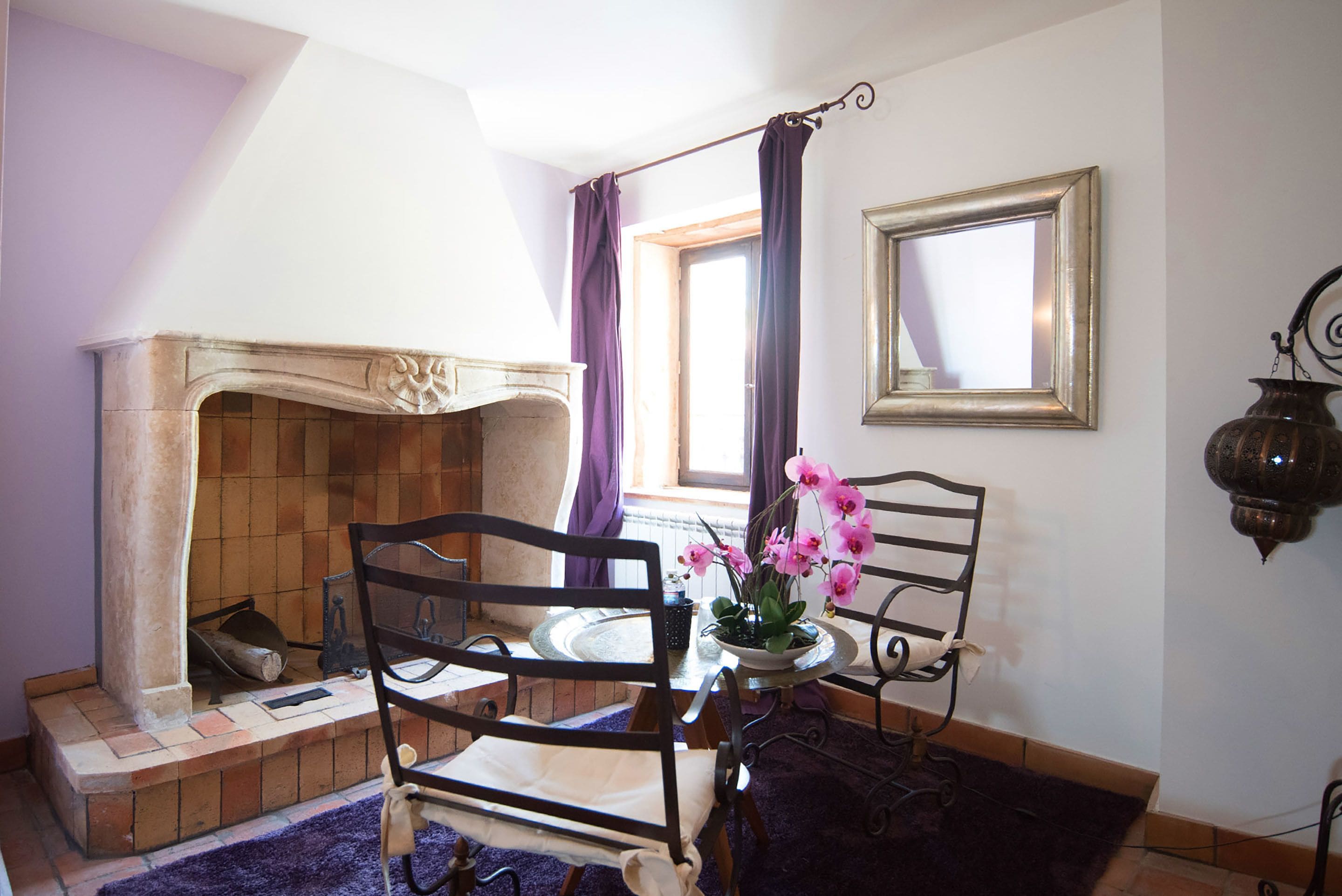 Ein Zimmer mit Kamin, zwei Stühlen, einem Tisch mit Blumenvase und einem fenster mit Vorhängen.
