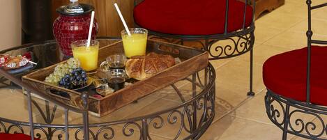 A breakfast tray with fruits, croissants, and orange juice on a glass table with red cushioned chairs.