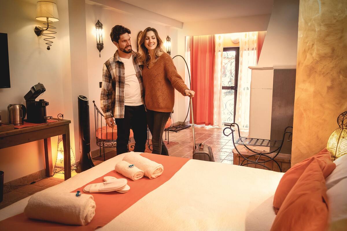 A hotel room with a bed, a table with towels, a suitcase, and a chair.