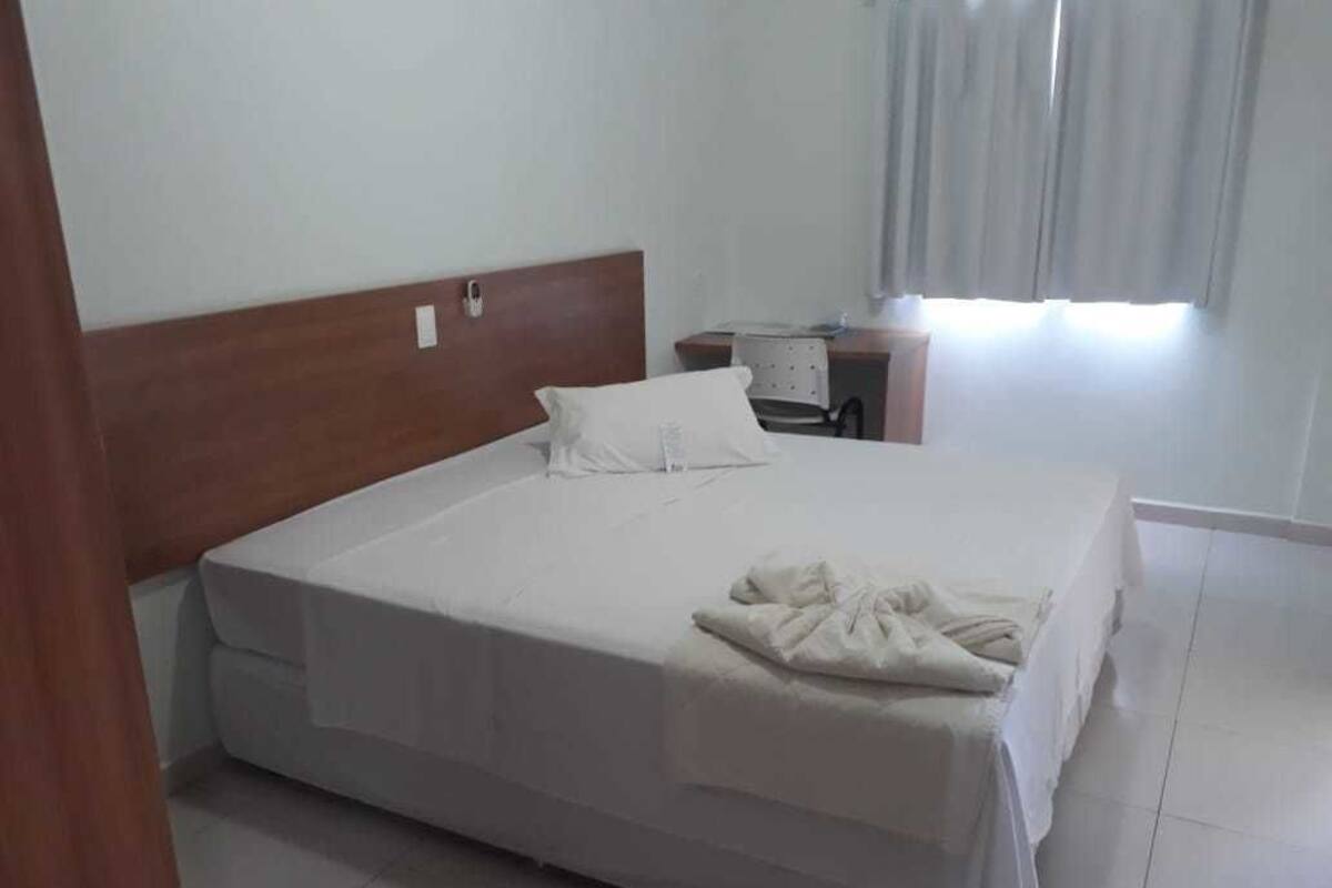 A neatly made bed with a white comforter and pillows, a wooden headboard, a small bedside table with a lamp, and a window with curtains.
