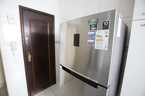 A stainless steel refrigerator with a digital display and various stickers, including a 10-year warranty and energy efficiency labels, in a kitchen with white tiles and a wooden door.