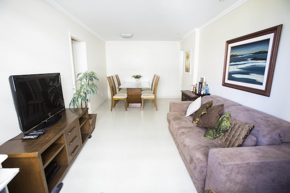 A living room with a flat-screen TV, wooden entertainment unit, a dining table with chairs, and a cushioned sofa.