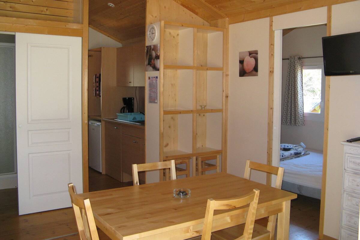 Ett matbord i trä med stolar, ett köksområde med skåp och ett kylskåp samt ett sovrum med en säng och gardiner.
