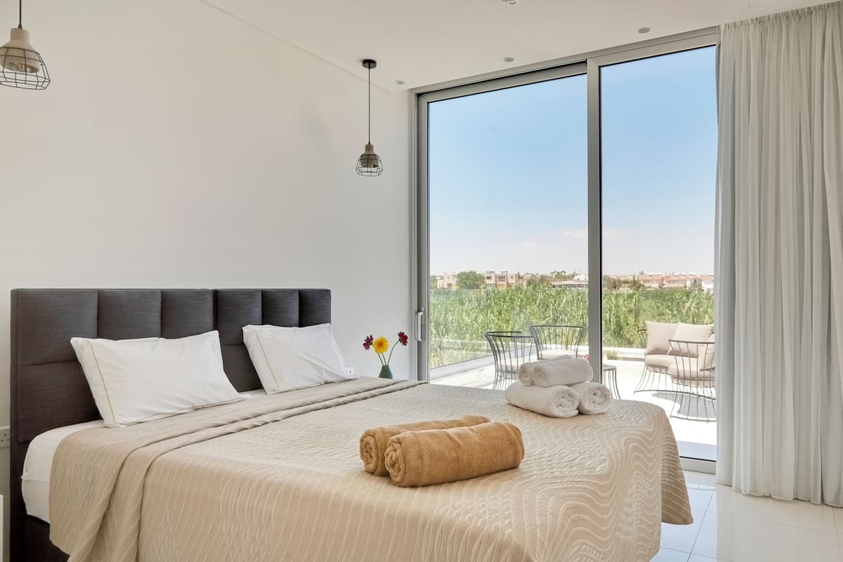 A modern hotel room with a large bed, a view of the outdoors, and a balcony with seating.