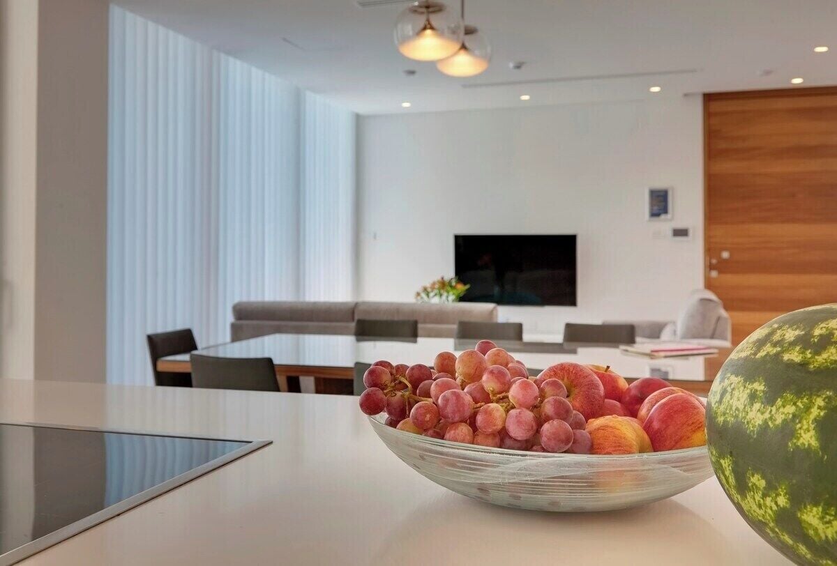 A modern dining area with a glass table, a bowl of fruit, and a large watermelon on the table.