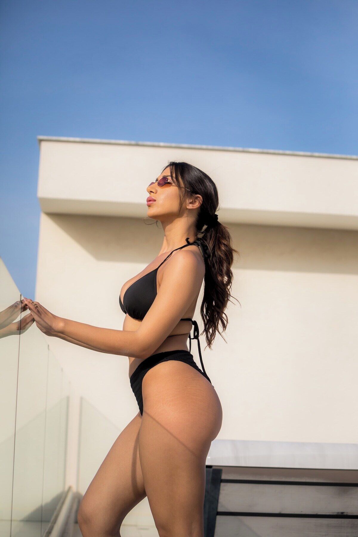 A person in a black bikini standing on a balcony with a clear sky in the background.