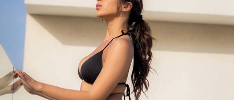 A person in a black bikini standing on a balcony with a clear sky in the background.