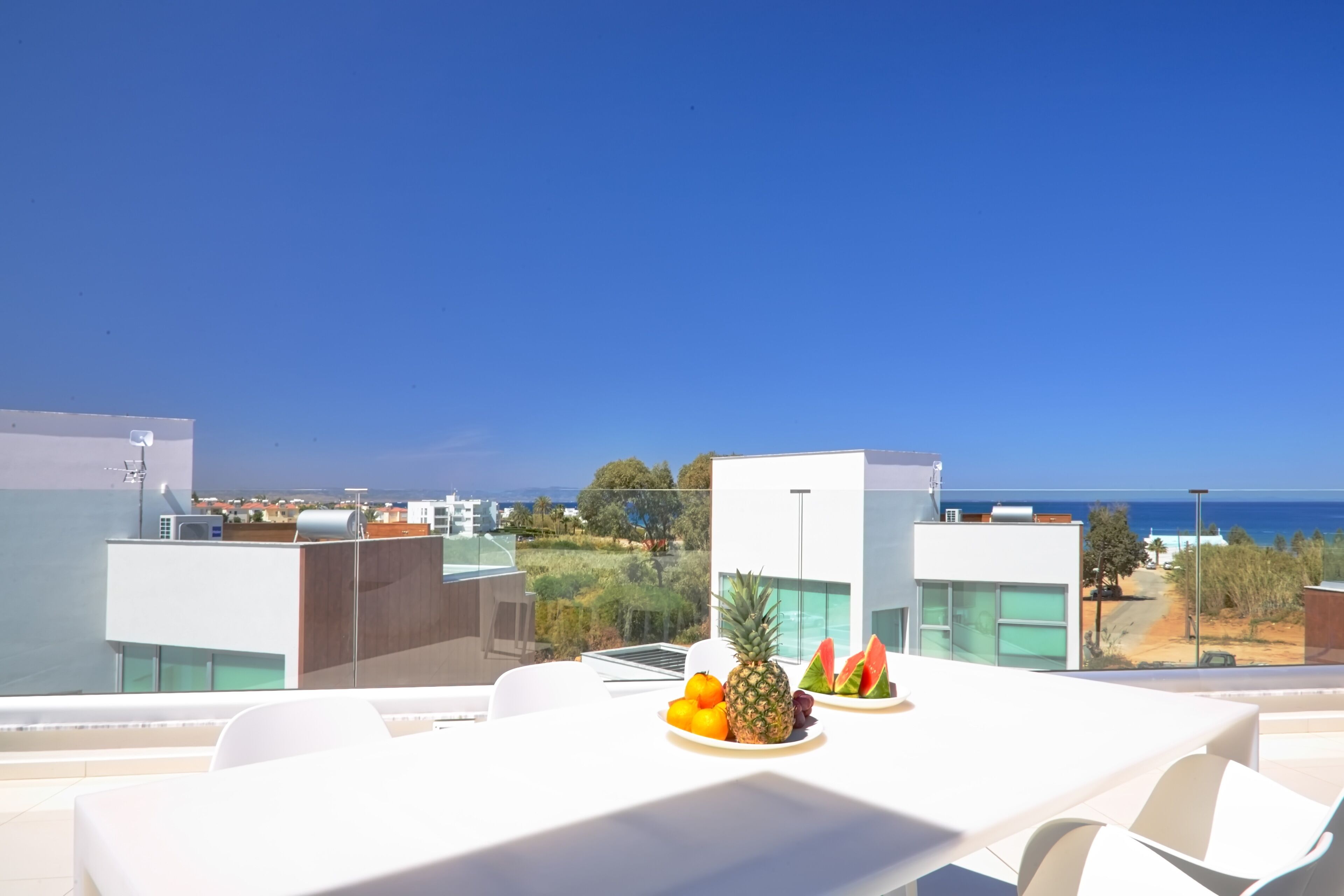 Eine Dachterrasse mit Esstisch, ausgestattet mit einer Obstschale mit Ananas, Orangen und Wassermelonenstücken. Die Terrasse bietet Blick auf ein modernes Gebäude und die Küste.