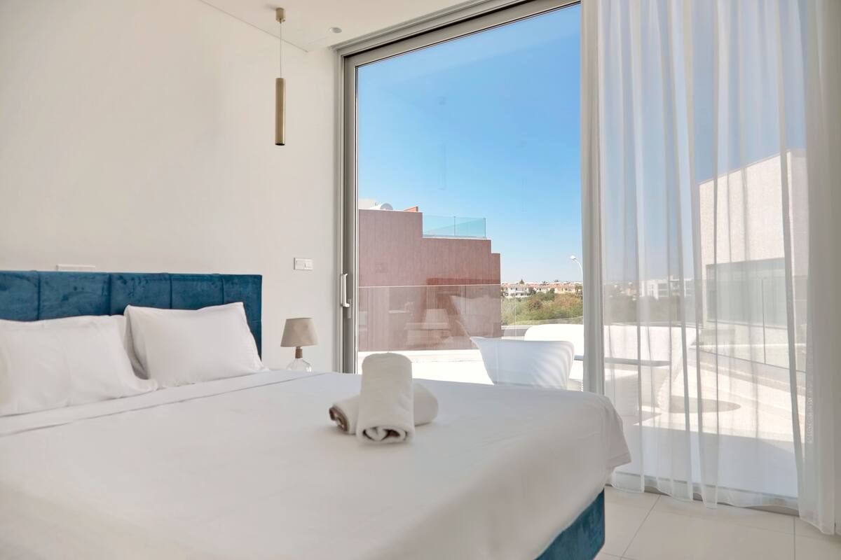 A hotel room with a large bed, a view through a glass door to the outside, and a blue headboard.