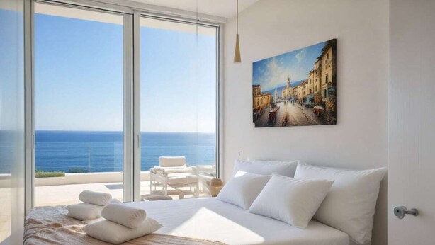 A bedroom with a large bed, white bedding, a painting of a street scene, and a view of the sea through floor-to-ceiling windows. - Sunrise Seaview Villas (Paralimni)
