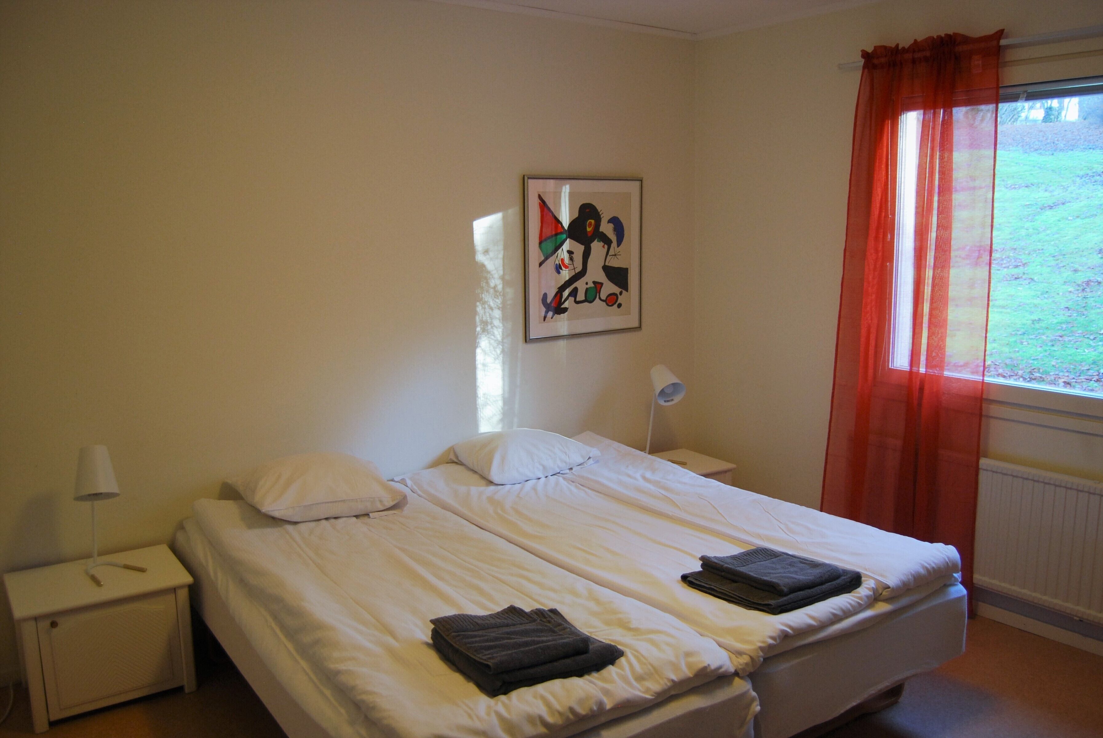 Una cama doble con ropa blanca, una mesita de noche con lámpara, un cuadro abstracto enmarcado en la pared y una ventana con cortinas rojas.