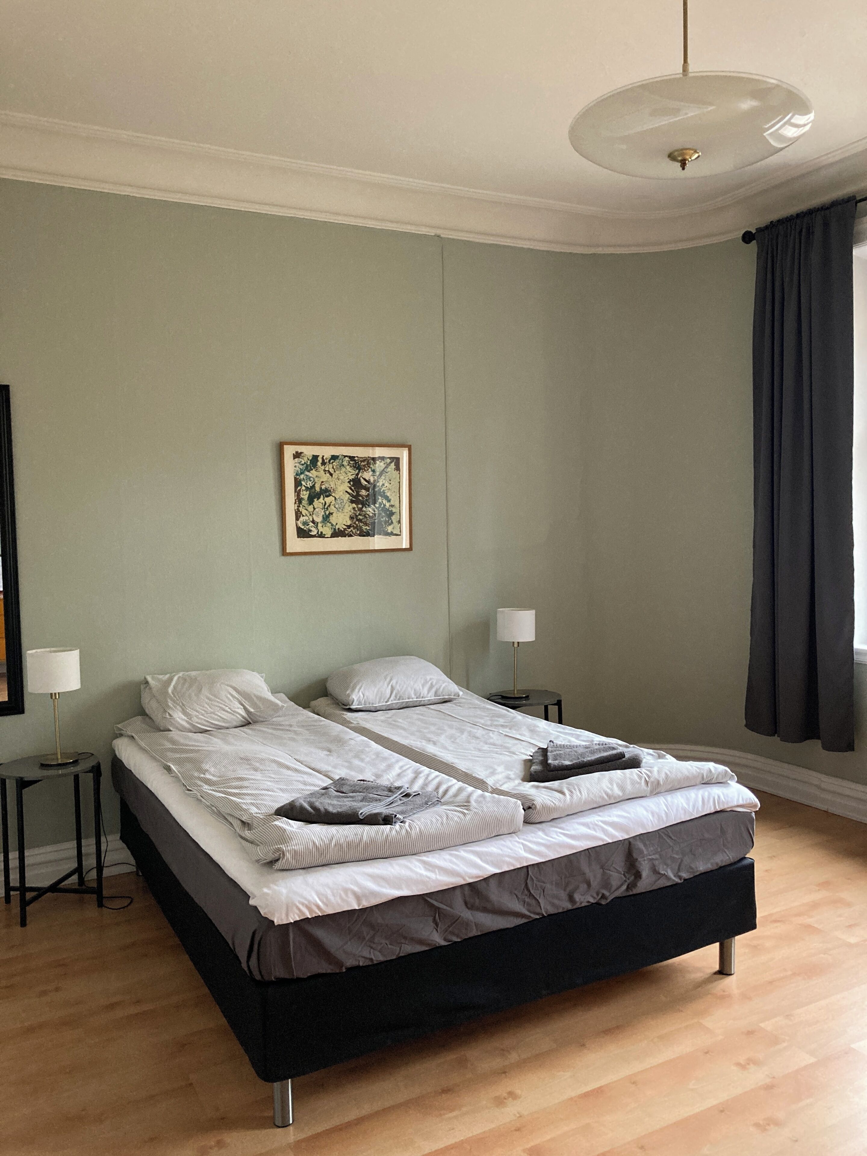 A double bed with a black frame and grey bedding, two bedside tables with lamps, a framed picture on the wall, and a window with dark curtains.