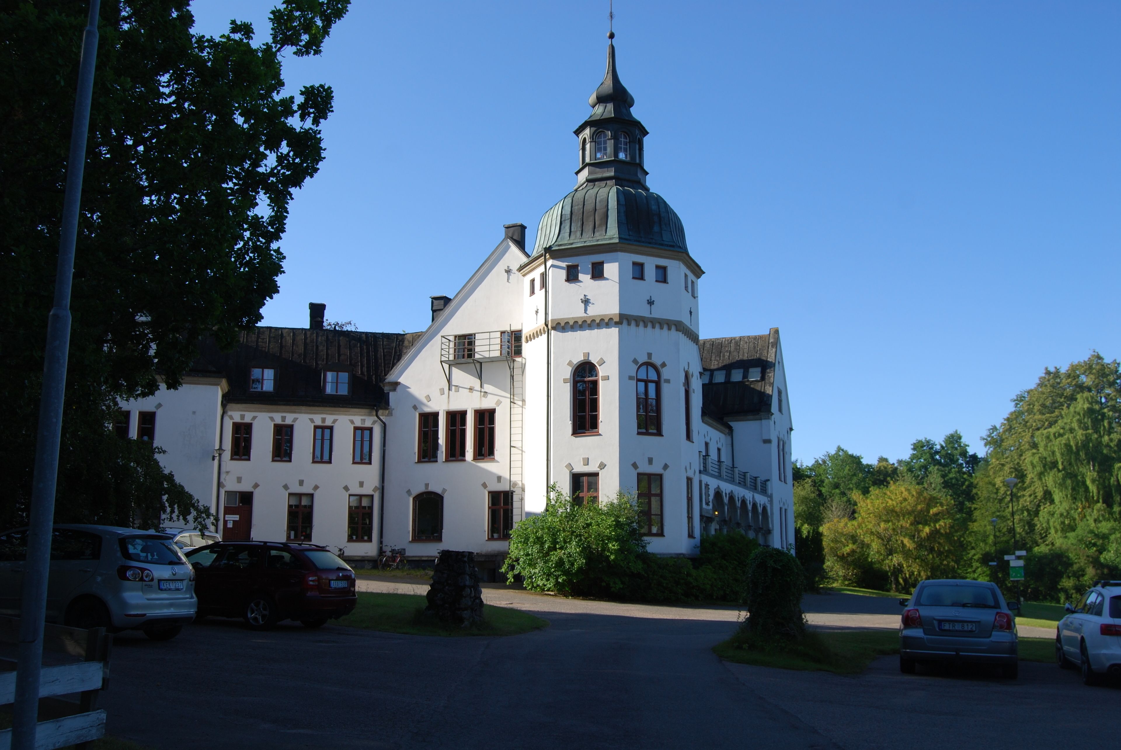 A white building with a prominent tower and a dome, surrounded by parked cars and greenery.