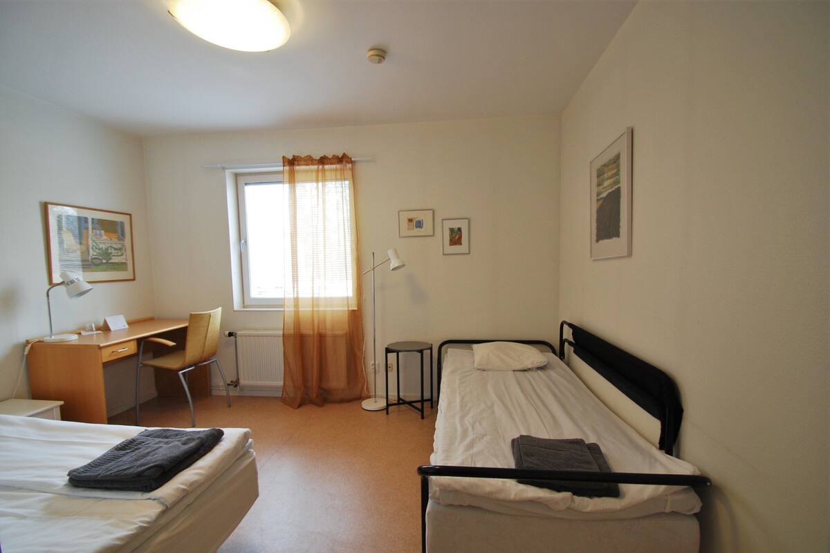 Una habitación pequeña y limpia con dos camas individuales, un escritorio, una silla y una ventana con cortinas.