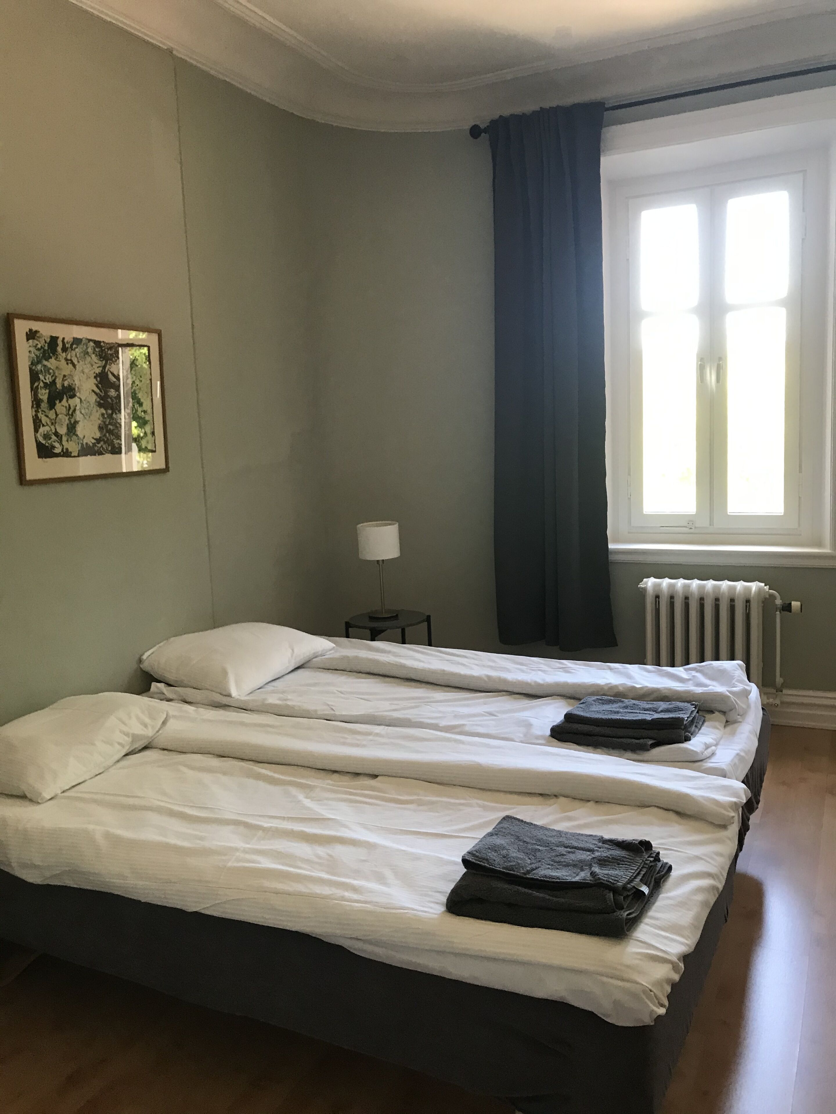 A room with two beds, a radiator, a window with curtains, and a framed picture on the wall.