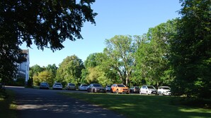 Un parking où plusieurs voitures sont garées en rangées, entouré d’arbres et d’un bâtiment doté d’une tour sur le côté gauche.