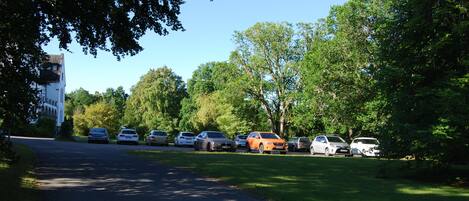 A parking lot with several cars parked in rows, surrounded by trees and a building with a tower on the left.