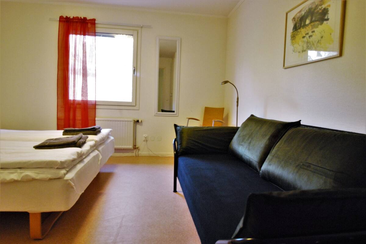 Una habitación con cama, sofá de color oscuro, ventana con cortinas rojas, espejo y lámpara.
