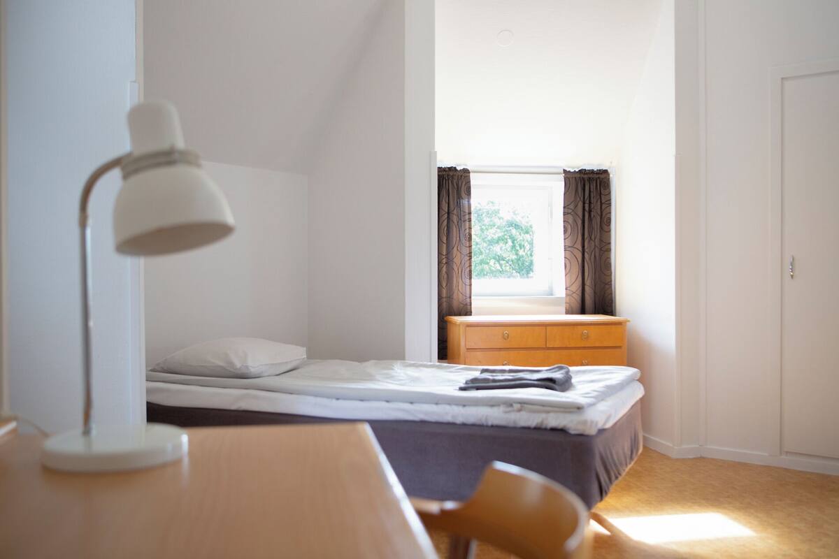 Un dormitorio pequeño y sencillo con una cama individual, un aparador de madera, un escritorio con lámpara y una ventana con cortinas.