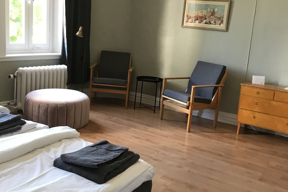 Un dormitorio con una cama, dos sillas, una mesita, un radiador y una ventana con cortinas.