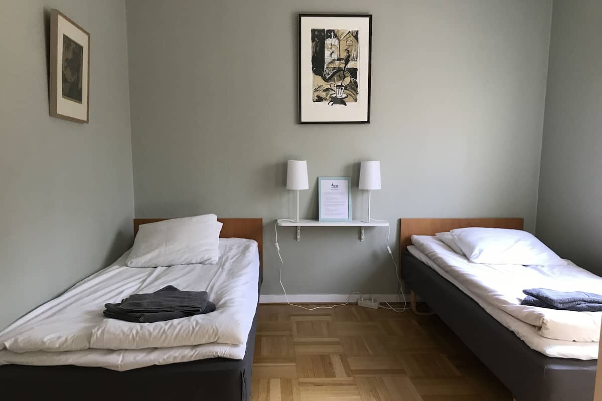 Habitación de hotel con dos camas individuales, suelo de madera, un cuadro en la pared y una mesita con dos lámparas.