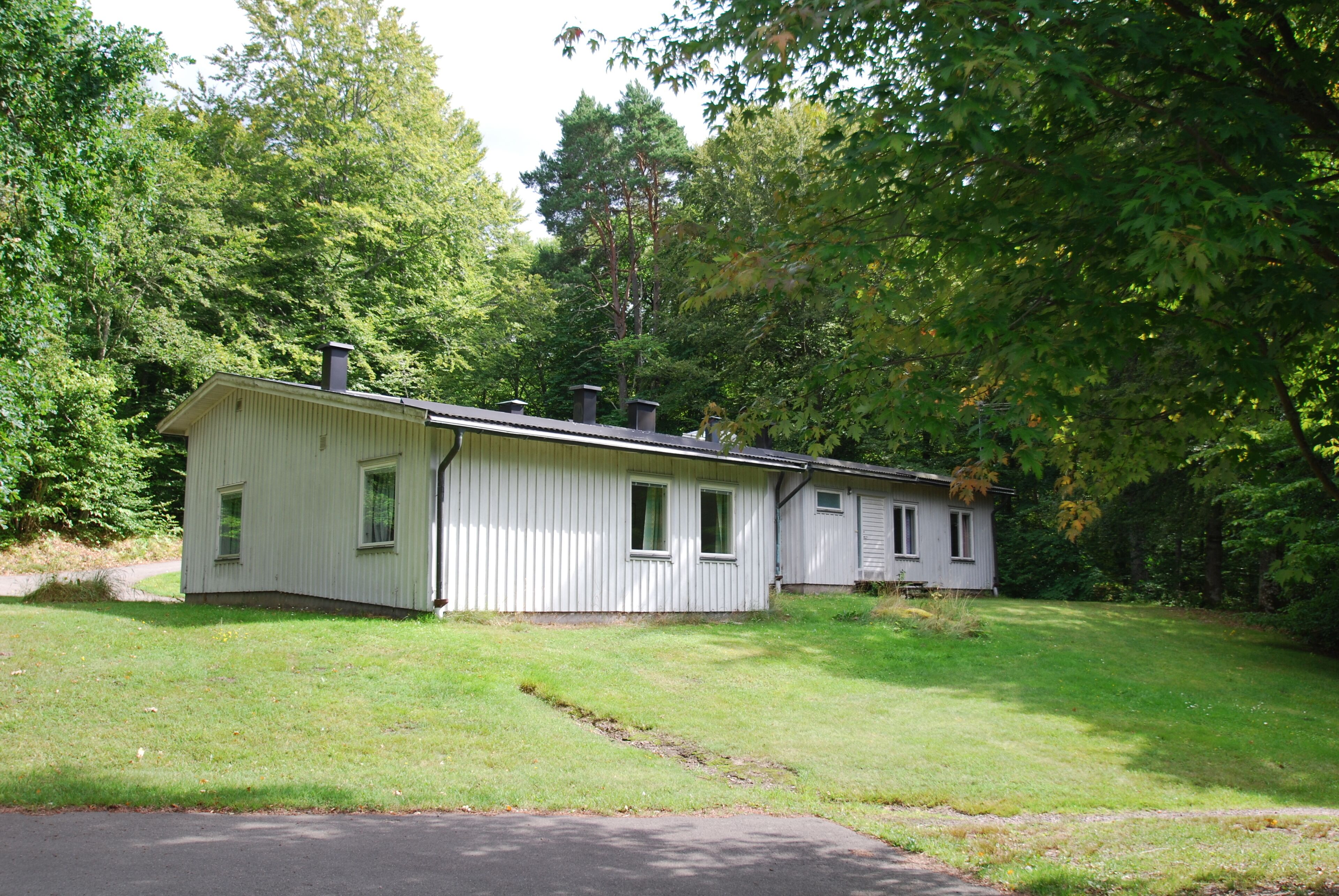 A single-story, white wooden cabin with a gabled roof, surrounded by lush green trees and a well-maintained lawn.