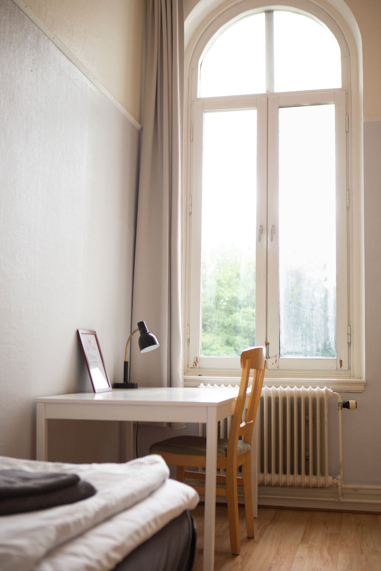 A room with a white desk, a chair, a bed, and a large window with curtains.