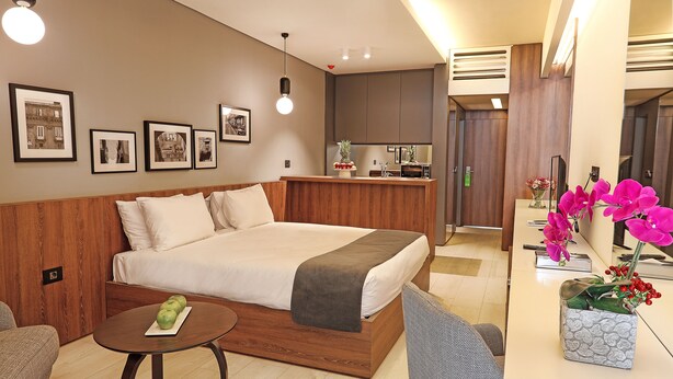 A modern hotel room with a large bed, a small table, a chair, and a kitchenette area. - Le Pave Residences (Ain Aar)