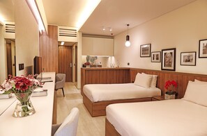 A hotel room with two beds, a desk with a vase of flowers, a chair, and a kitchenette in the background. - Le Pave Residences (Ain Aar)