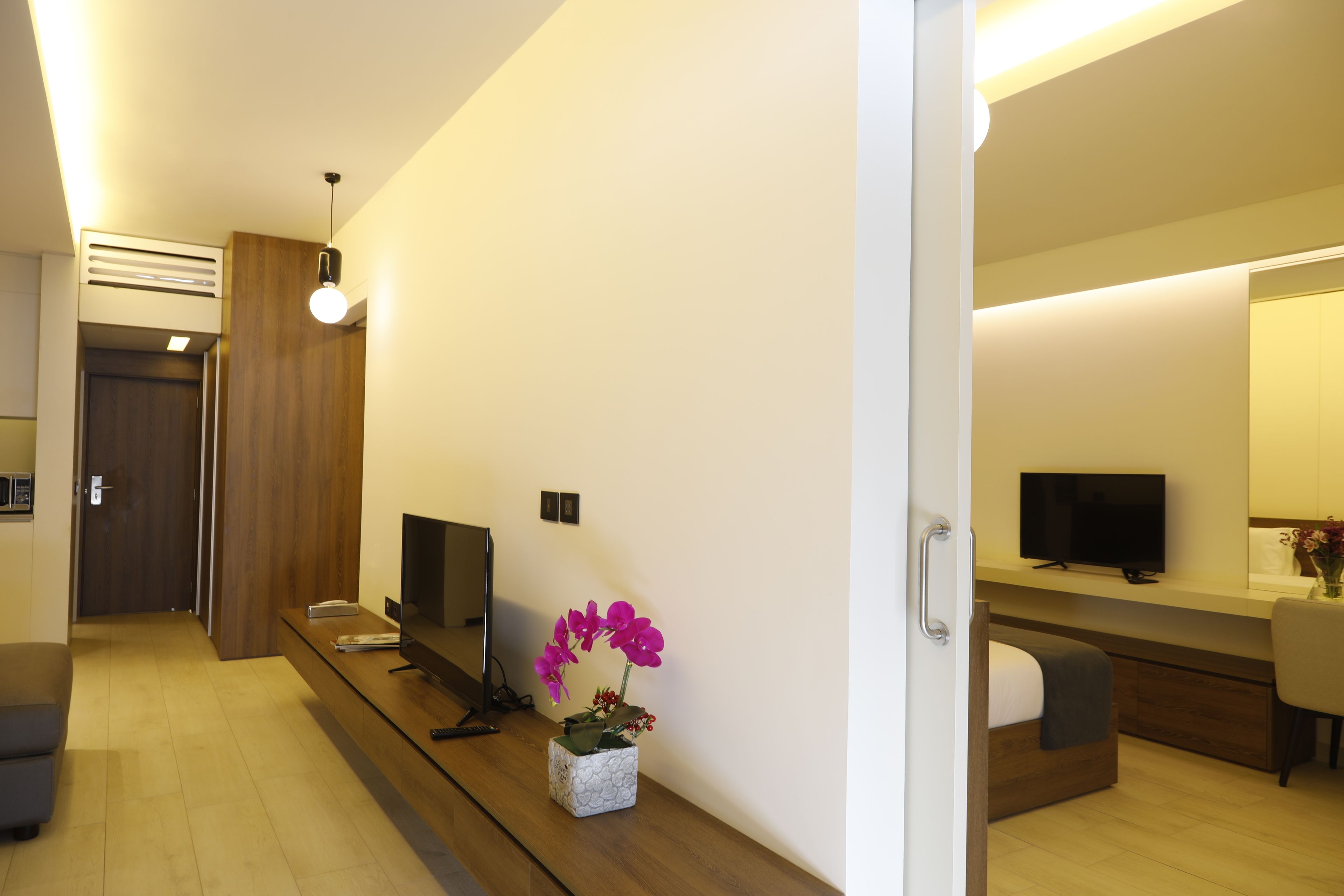 A modern hotel room with a wooden floor, a flat-screen TV, a small table with a vase of flowers, and a large mirror above the bed.