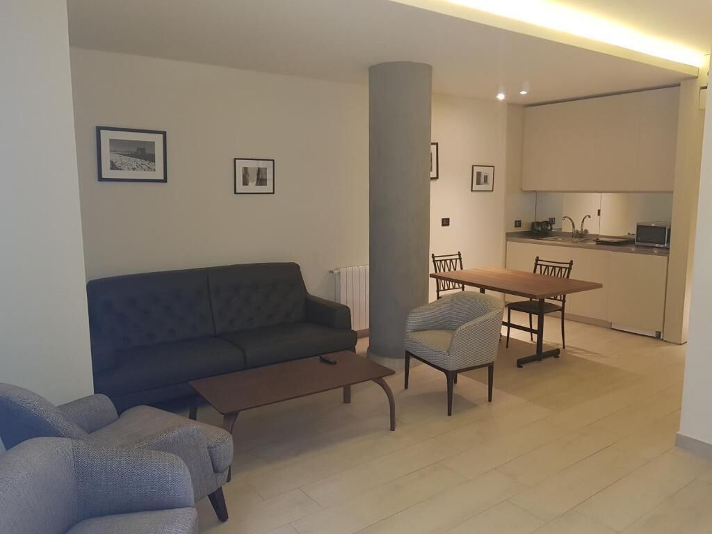 Ein modernes Wohnzimmer mit einer Couch, einem Sessel und einem Esstisch mit Stühlen. Im Hintergrund ist eine Küchenzeile erkennbar.