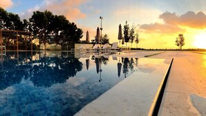 A swimming pool with lounge chairs and umbrellas, surrounded by trees and a sunset. - Le Pave Residences (Ain Aar)