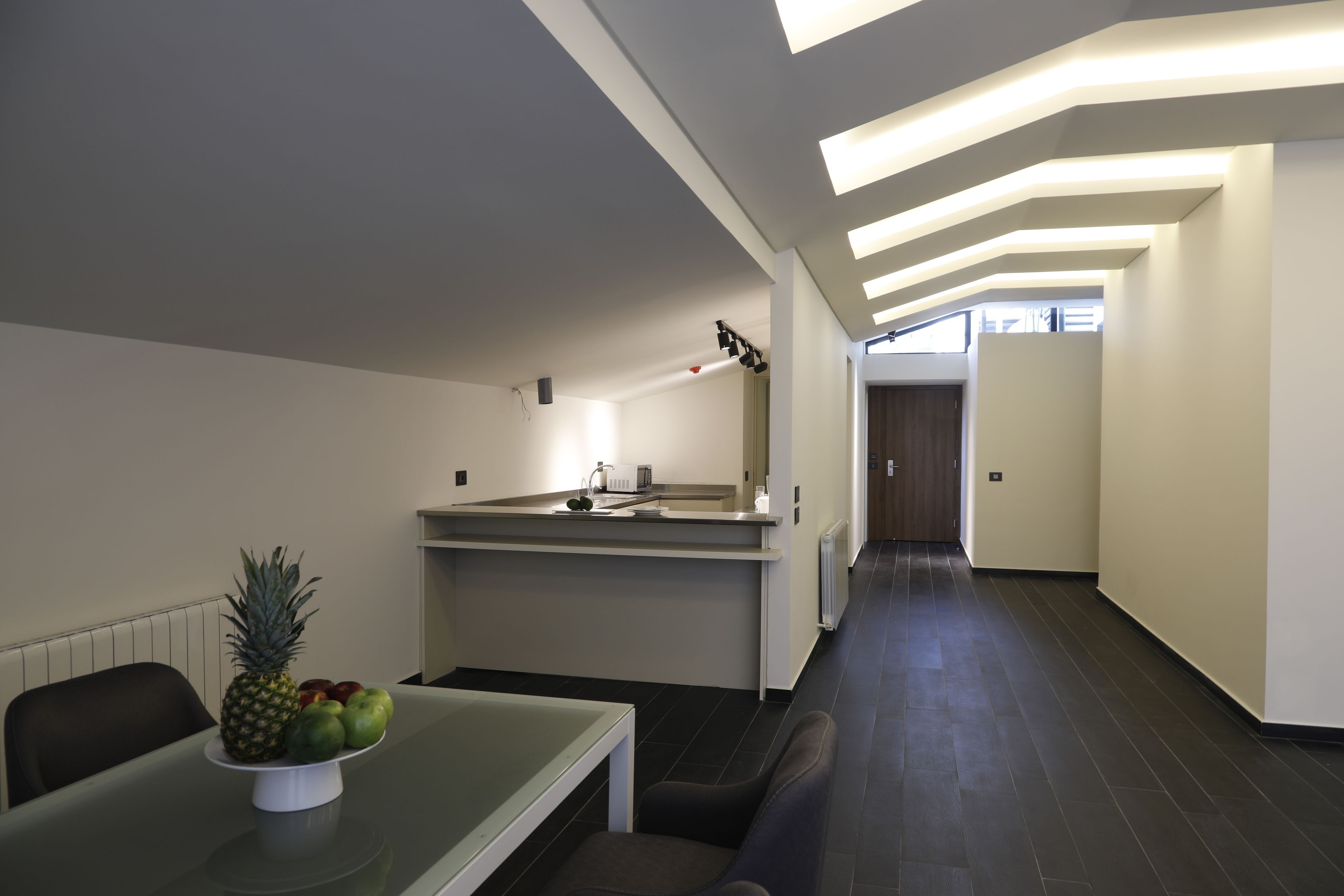 A modern kitchen with a dining area, featuring a long countertop, a bowl of fruit, and a seating arrangement.