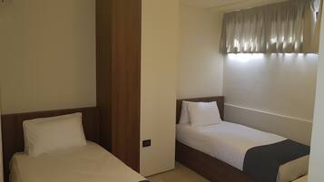 A hotel room with two single beds, a wooden wardrobe, a bedside table, and a window with curtains.